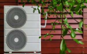 outdoor heat pump unit against red siding wall with some foliage in the foreground