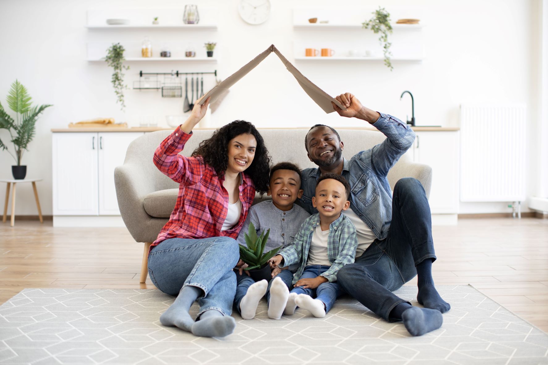 family of four sitting down together in their living room; parents on the ends are holding up a triangle of cardboard to emulate a home over their heads