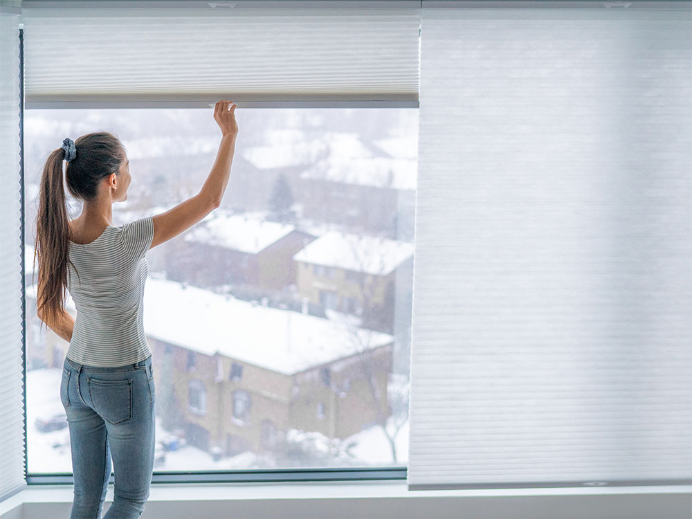 A woman lowers cellular shades. A winter scene is visible through the window.