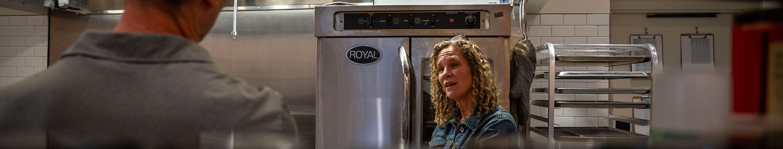 A woman stands next to an electric oven in a commercial kitchen. She is talking to a man whose upper back and head is visible in the foreground.
