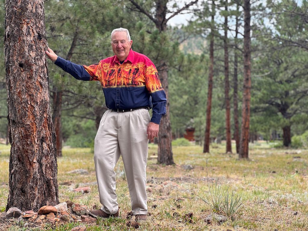 John Miller stands by a ponderosa pine tree in a wooded forest with green trees and grass.